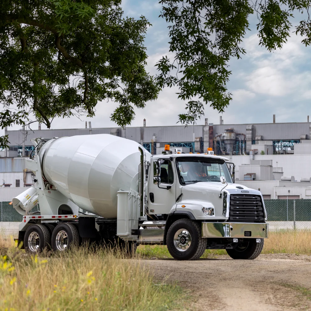 Freightliner cement mixer truck parked near trees with an industrial backdrop